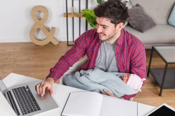 Padre che tiene in braccio il figlio mentre studia al computer, simbolo di crescita professionale e equilibrio familiare.